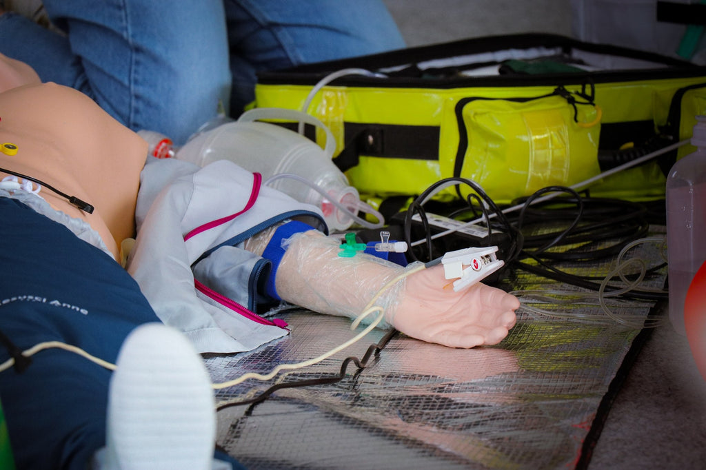 ACLS Refresh: intravenöser Zugang am Arm des Simulators mit Notfallrucksack im Hintergrund