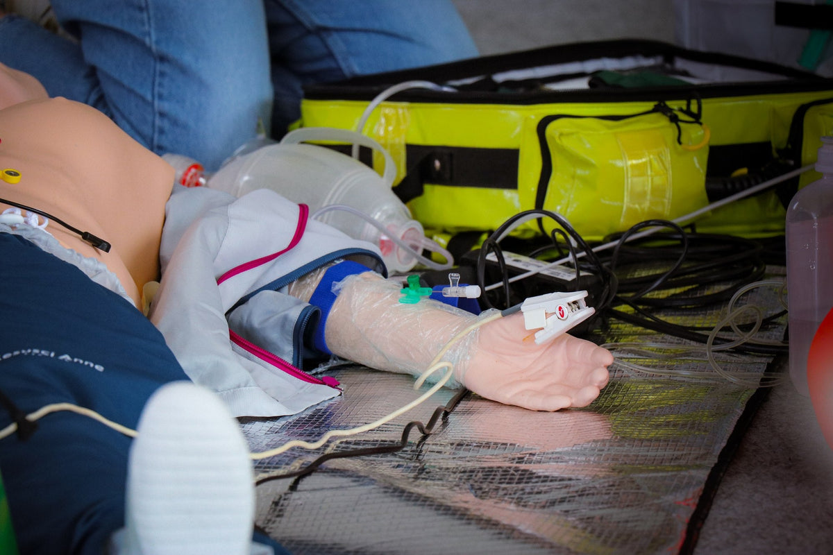 ACLS Refresh: intravenöser Zugang am Arm des Simulators mit Notfallrucksack im Hintergrund
